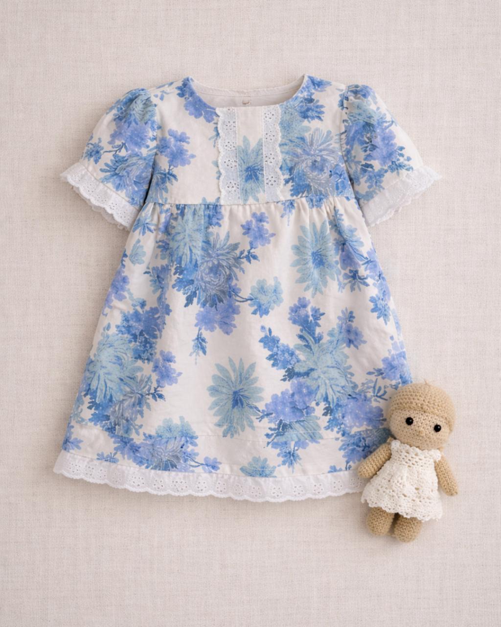 A flat lay photograph of the Alma baby dress, featuring a white fabric with a soft blue floral and leaf print. The dress has short puffed sleeves and is detailed with white eyelet lace trim on the chest, sleeve cuffs, and hem. It is styled on a neutral linen background next to a small, handmade tan crochet doll wearing a white lace dress.