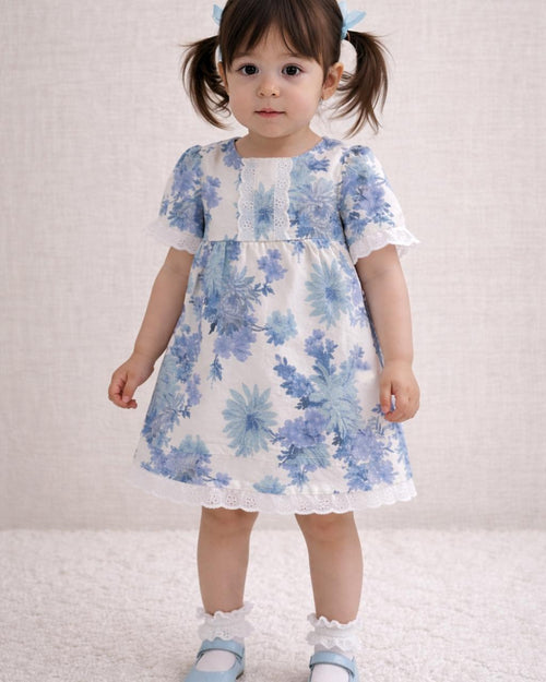 A lifestyle photograph of a young girl with pigtails standing on a soft white rug. She is wearing the Alma dress, a white short-sleeved garment featuring a blue floral print and white eyelet lace trim on the neckline, sleeves, and hem. The look is completed with white ruffle-top socks and light blue Mary Jane shoes against a neutral, textured wall.
