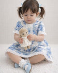 A lifestyle photograph of a young girl with pigtails sitting on a soft white rug. She is wearing the Alma dress, which features a blue and white floral toile print, short ruffled sleeves, and delicate white eyelet lace trim along the neckline, sleeves, and hem. She is holding a small, handmade crochet doll, and wearing white ruffle socks with light blue Mary Jane shoes.
