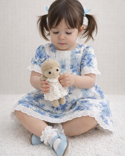 A lifestyle photograph of a young girl with pigtails sitting on a soft white rug. She is wearing the Alma dress, which features a blue and white floral toile print, short ruffled sleeves, and delicate white eyelet lace trim along the neckline, sleeves, and hem. She is holding a small, handmade crochet doll, and wearing white ruffle socks with light blue Mary Jane shoes.