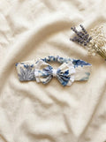 A flat lay photograph of the Alma baby headband, featuring a soft cream fabric with a blue and grey vintage-style floral pattern. The headband has an elasticated back for a comfortable fit and is topped with a matching floral bow that is layered over a white ruffled fabric. It is styled on a neutral, draped linen background next to a sprig of dried lavender and white baby's breath flowers.