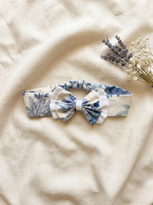 A flat lay photograph of the Alma baby headband, featuring a soft cream fabric with a blue and grey vintage-style floral pattern. The headband has an elasticated back for a comfortable fit and is topped with a matching floral bow that is layered over a white ruffled fabric. It is styled on a neutral, draped linen background next to a sprig of dried lavender and white baby's breath flowers.