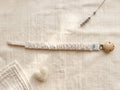 A flat lay photograph of a pacifier clip featuring a fabric strap with a thin beige and cream horizontal stripe pattern. The strap has a gathered, ruffled texture and is secured to a round, natural-finish wooden clip. It is styled on a textured cream muslin background with a small white felt heart and a single sprig of dried lavender.