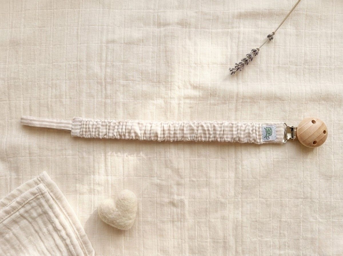A flat lay photograph of a pacifier clip featuring a fabric strap with a thin beige and cream horizontal stripe pattern. The strap has a gathered, ruffled texture and is secured to a round, natural-finish wooden clip. It is styled on a textured cream muslin background with a small white felt heart and a single sprig of dried lavender.
