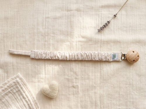 A flat lay photograph of a pacifier clip featuring a fabric strap with a thin beige and cream horizontal stripe pattern. The strap has a gathered, ruffled texture and is secured to a round, natural-finish wooden clip. It is styled on a textured cream muslin background with a small white felt heart and a single sprig of dried lavender.
