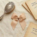 A flat lay photograph of a large, perfectly tied beige grosgrain ribbon hair bow with long tails. The bow is resting on a neutral linen fabric, surrounded by vintage aesthetic props, including an antique silver hairbrush and old books.