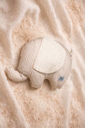 A top-down photograph of a small elephant-shaped plush cushion featuring a beige and white vertical stripe pattern. The elephant has a large, textured cream-colored ear and a small grey brand label near the back. It is styled on a soft cream fabric backdrop over a layer of fine sand, creating a warm, natural nursery aesthetic.