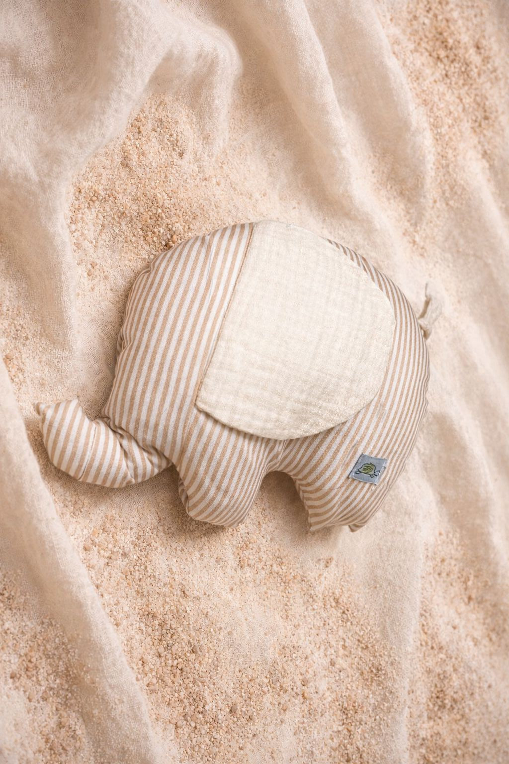 A top-down photograph of a small elephant-shaped plush cushion featuring a beige and white vertical stripe pattern. The elephant has a large, textured cream-colored ear and a small grey brand label near the back. It is styled on a soft cream fabric backdrop over a layer of fine sand, creating a warm, natural nursery aesthetic.