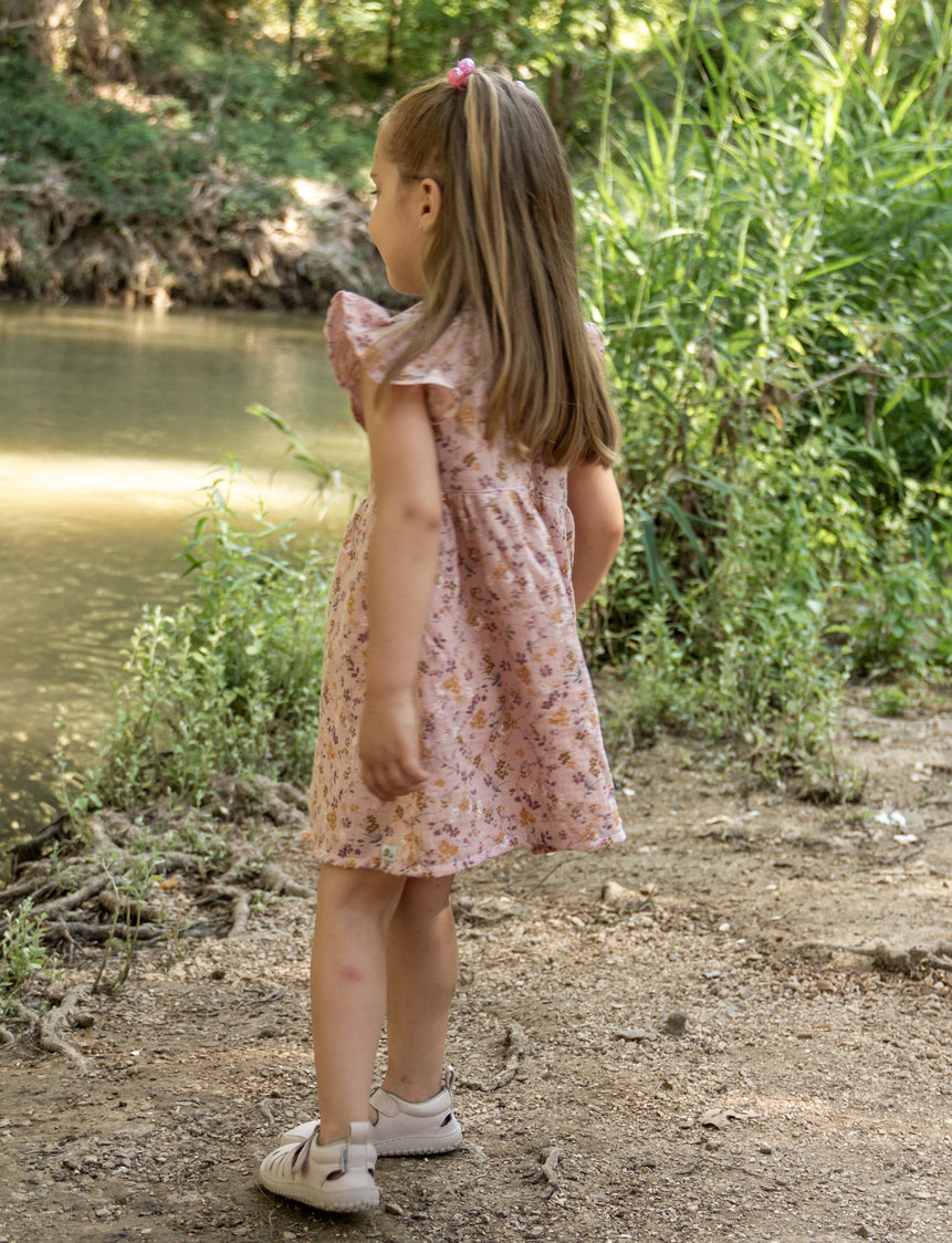 A three-quarter back view of a young girl standing outdoors near a riverbank, wearing the light pink Bella dress with flutter sleeves and a subtle all-over floral print. The girl is walking away from the camera, her long blonde hair visible, with lush green foliage and a calm river filling the background.
