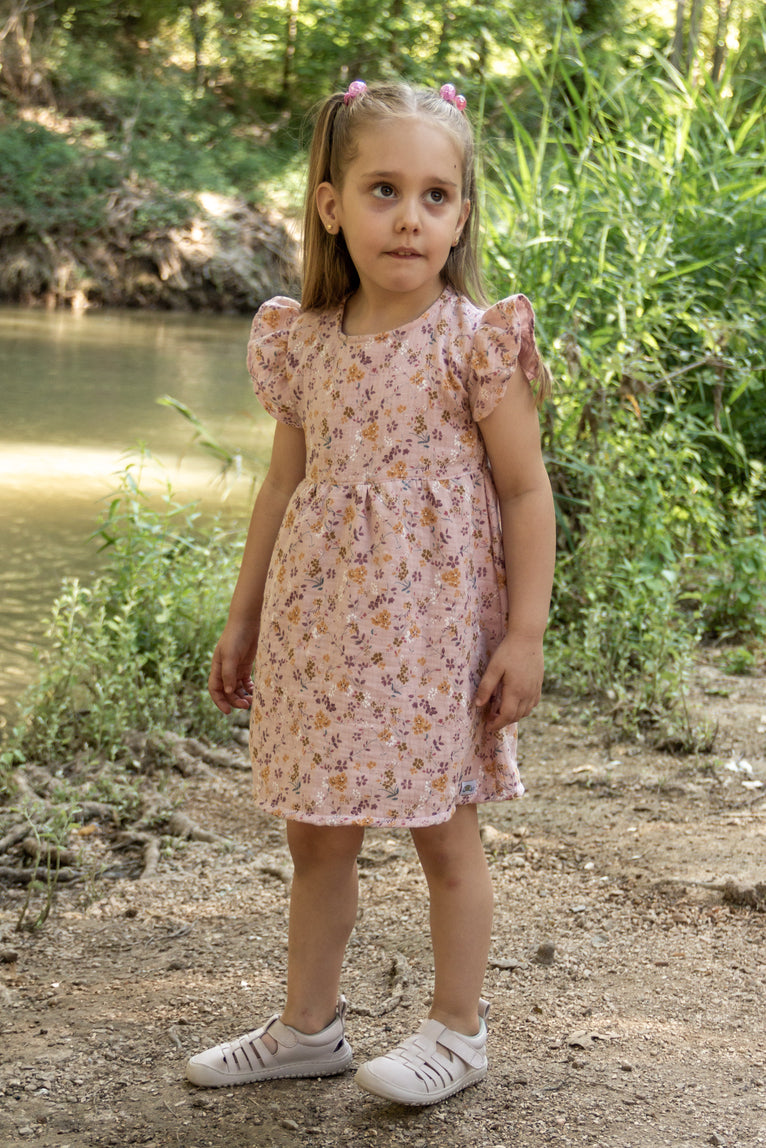 A full-length lifestyle photograph of a young girl standing outdoors on a dirt path, wearing the pink floral Bella dress with flutter sleeves and a flared skirt. The fabric is light pink with small orange and purple floral clusters, and the girl has blonde pigtails and white shoes, standing near a river with lush trees and foliage