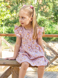 A lifestyle photograph of a young girl sitting on a wooden fence outdoors, wearing the Bella pink floral dress with flutter cap sleeves and a flared skirt. The fabric is patterned with small mustard yellow and purple floral sprigs, set against a sunlit garden background with lush greenery.