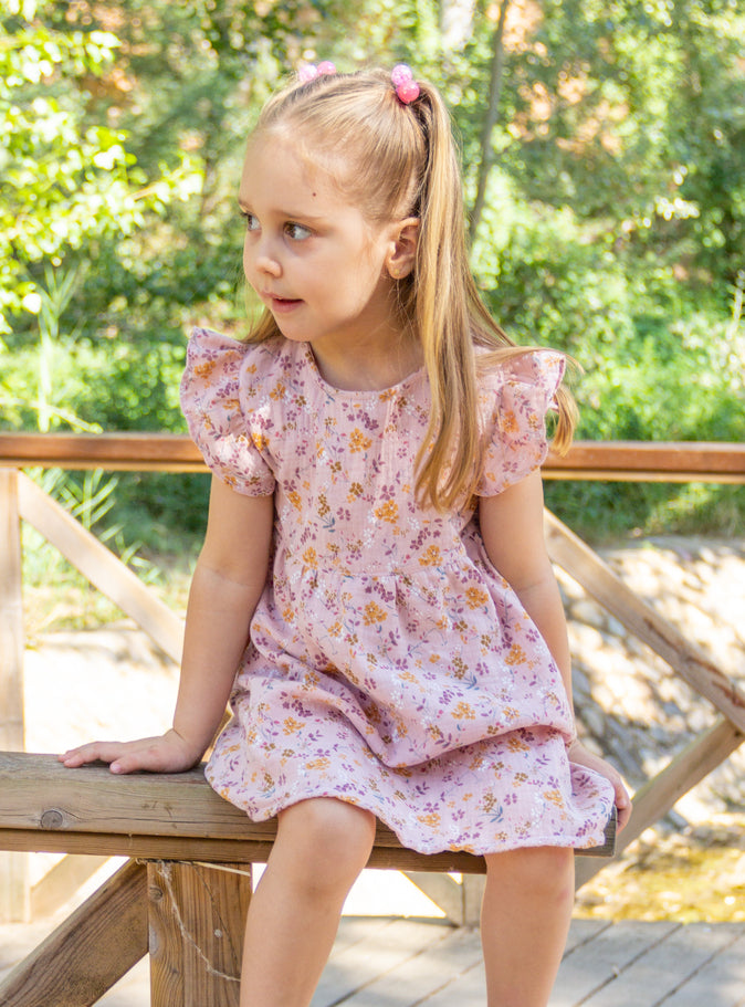 A lifestyle photograph of a young girl sitting on a wooden fence outdoors, wearing the Bella pink floral dress with flutter cap sleeves and a flared skirt. The fabric is patterned with small mustard yellow and purple floral sprigs, set against a sunlit garden background with lush greenery.