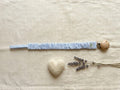 A flat lay photograph of a pacifier clip featuring a fabric strap with a thin light blue and white vertical stripe pattern. The strap is gathered with a ruffled, elasticated texture and is attached to a round, natural light-wood clip. It is styled on a neutral, textured cream fabric background alongside a small white felt heart and sprigs of dried lavender.
