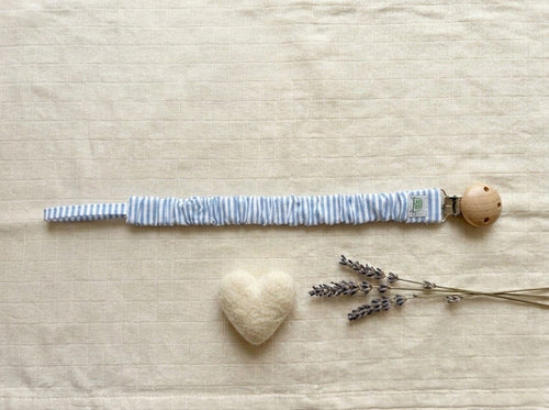 A flat lay photograph of a pacifier clip featuring a fabric strap with a thin light blue and white vertical stripe pattern. The strap is gathered with a ruffled, elasticated texture and is attached to a round, natural light-wood clip. It is styled on a neutral, textured cream fabric background alongside a small white felt heart and sprigs of dried lavender.