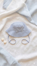 A flat lay photograph of a handmade baby bucket hat featuring blue and white horizontal stripes. The hat includes matching fabric chin straps and a small green turtle brand label on the brim. It is styled on a neutral linen backdrop alongside a sprig of dried lavender and a small smooth stone in soft, natural light.