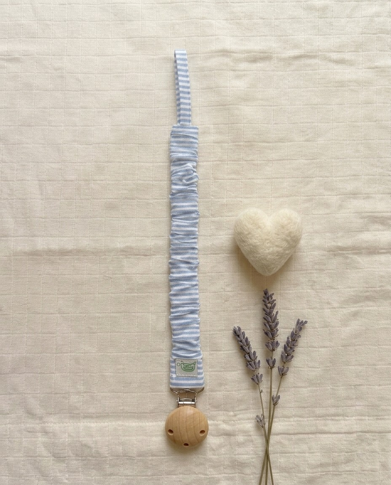 A product flat-lay photograph of a baby pacifier clip with light blue and white horizontal stripes lying vertically on a soft cream linen backdrop. The pacifier clip features a gathered fabric sleeve, a loop at the top, a small woven brand label near the base, and a round natural wooden clip at the bottom. A small plush white heart and a few sprigs of dried lavender accent the right side.