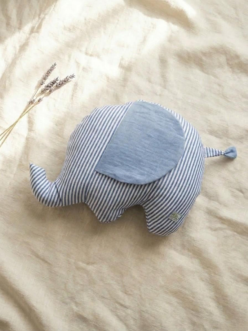 A handmade elephant-shaped plush cushion featuring a blue and white vertical striped body and a solid light blue muslin fabric ear. The cushion is displayed on a neutral linen backdrop with a sprig of dried lavender in the upper left corner, creating a soft and calm nursery aesthetic.