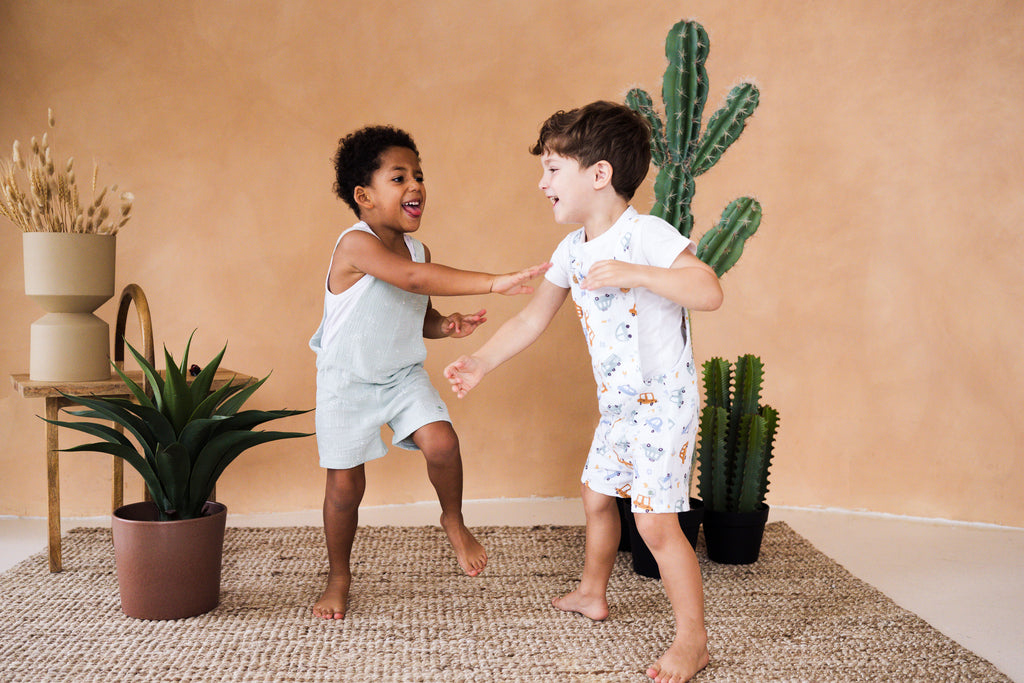 A dynamic lifestyle photograph of two young boys playing and laughing barefoot on a woven jute rug inside a studio with a warm terracotta-colored wall. The boy on the left is wearing a light mint green bubble-style romper featuring a subtle white giraffe and elephant line-art print, worn over a white tank top. The boy on the right is dancing in a white short romper decorated with a colorful pattern of cars and trucks, styled over a simple white t-shirt. The scene is decorated with a tall potted cactus.