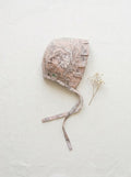 A flat lay photograph of the Carmen baby bonnet, featuring a neutral beige fabric with a dark brown vintage toile floral pattern. The bonnet has a ruffled brim, matching chin straps, and a small green brand label. It is styled on a crisp white linen background next to a delicate sprig of dried white baby's breath flowers.