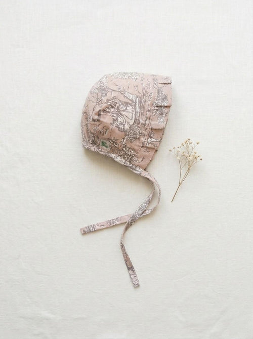 A flat lay photograph of the Carmen baby bonnet, featuring a neutral beige fabric with a dark brown vintage toile floral pattern. The bonnet has a ruffled brim, matching chin straps, and a small green brand label. It is styled on a crisp white linen background next to a delicate sprig of dried white baby's breath flowers.
