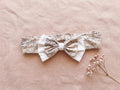 A flat lay photograph of the Carmen baby headband, featuring a beige fabric with a delicate brown vintage floral or toile-style pattern. The headband includes an elasticated back and is adorned with a layered bow—a smaller floral bow set atop a larger white fabric bow. It is styled against a soft, dusty pink linen backdrop next to a sprig of dried pink baby's breath flowers.