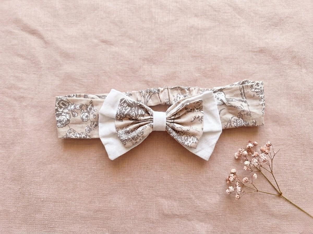 A flat lay photograph of the Carmen baby headband, featuring a beige fabric with a delicate brown vintage floral or toile-style pattern. The headband includes an elasticated back and is adorned with a layered bow—a smaller floral bow set atop a larger white fabric bow. It is styled against a soft, dusty pink linen backdrop next to a sprig of dried pink baby's breath flowers.