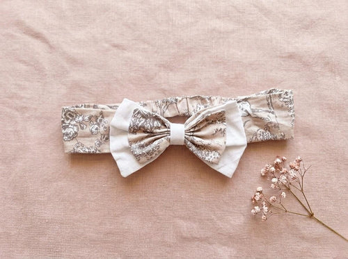A flat lay photograph of the Carmen baby headband, featuring a beige fabric with a delicate brown vintage floral or toile-style pattern. The headband includes an elasticated back and is adorned with a layered bow—a smaller floral bow set atop a larger white fabric bow. It is styled against a soft, dusty pink linen backdrop next to a sprig of dried pink baby's breath flowers.
