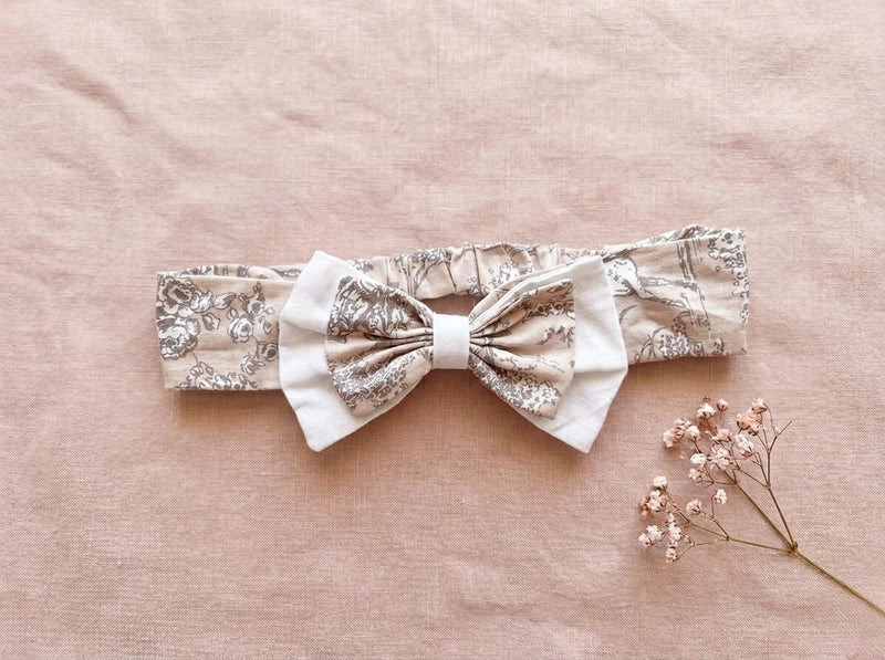 A flat lay photograph of the Carmen baby headband, featuring a beige fabric with a delicate brown vintage floral or toile-style pattern. The headband includes an elasticated back and is adorned with a layered bow—a smaller floral bow set atop a larger white fabric bow. It is styled against a soft, dusty pink linen backdrop next to a sprig of dried pink baby's breath flowers.