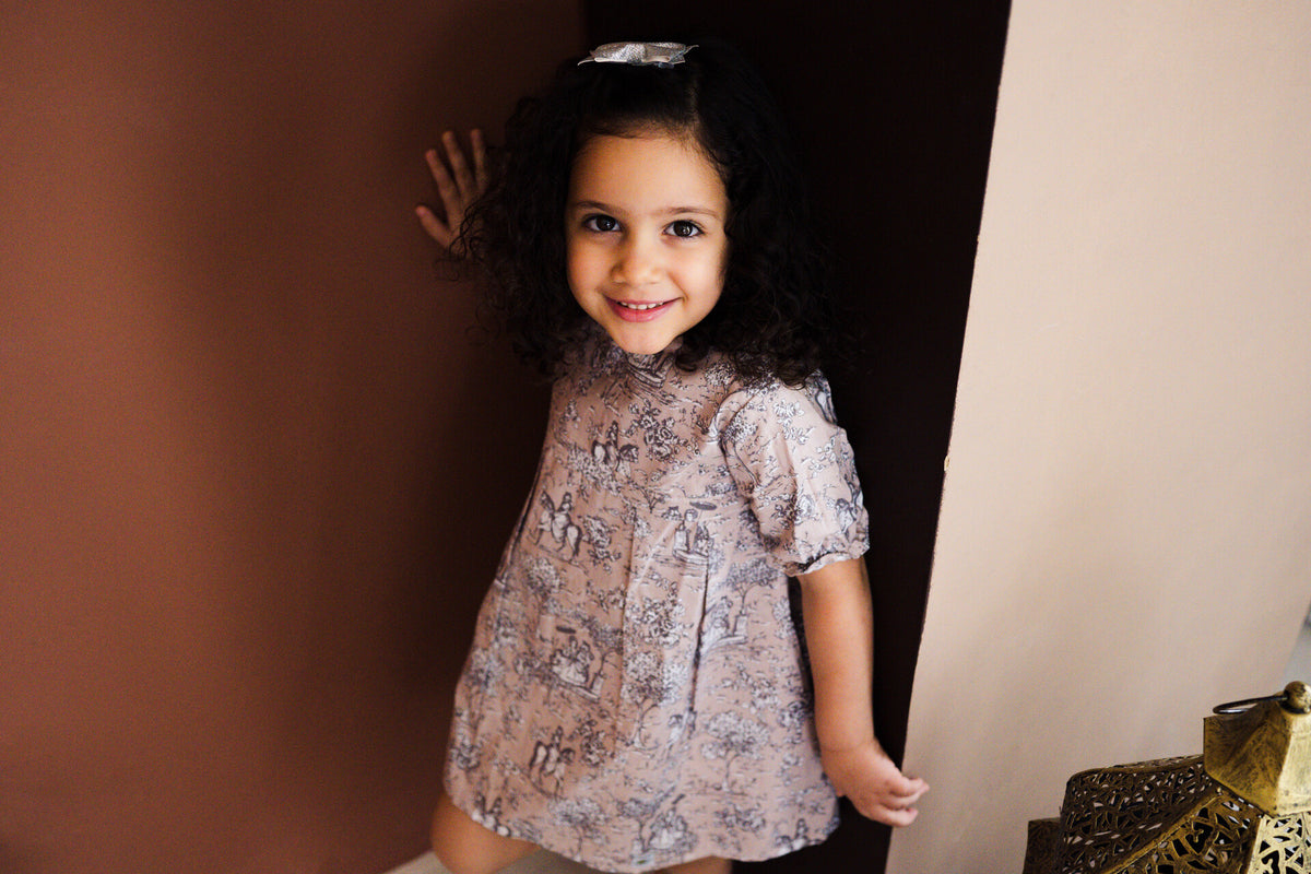 A close-up studio portrait of a smiling A close-up studio portrait of a smiling young girl with dark curly hair and a hair clip, wearing the Carmen short-sleeved dress with a classic grey and white toile de Jouy pattern on a light beige fabric. She peeks around a brown partition wall in a warm, minimalist indoor set.