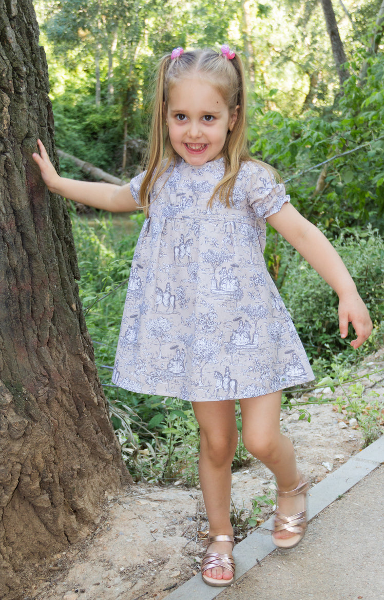 A full-length outdoor lifestyle photograph of a smiling young girl leaning against a large tree trunk, wearing the Carmen short-sleeved A-line dress featuring a classic grey and white toile de Jouy pattern on a light beige fabric. She has blonde pigtails and metallic strappy sandals, set against a lush, sunlit park background.