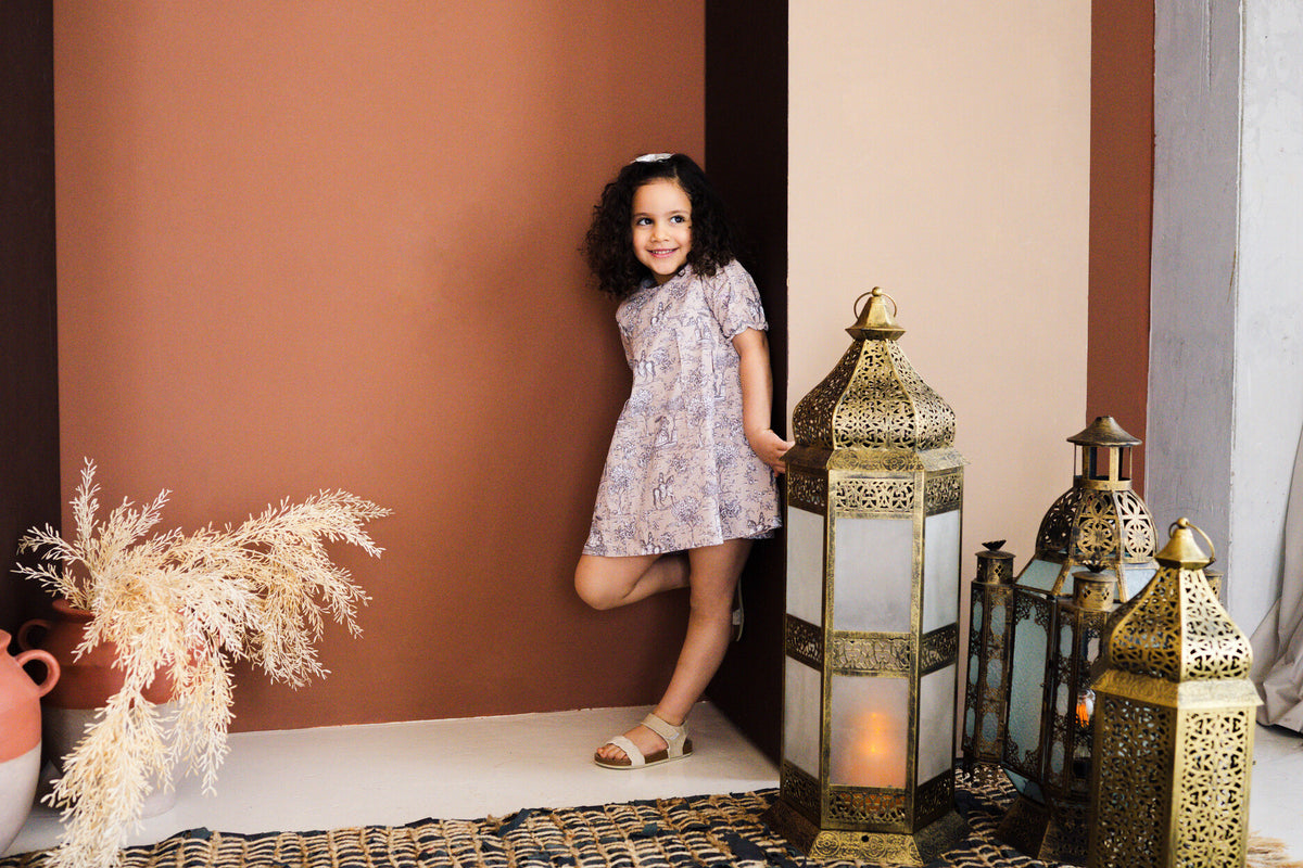 A full-length studio lifestyle photograph of a smiling young girl leaning against a brown accent wall, wearing the Carmen short-sleeved dress with a grey and white toile de Jouy pattern on beige fabric. She wears a hair clip and beige sandals while posing on a woven rug surrounded by ornate brass lanterns and dried pampas grass in terracotta vases.