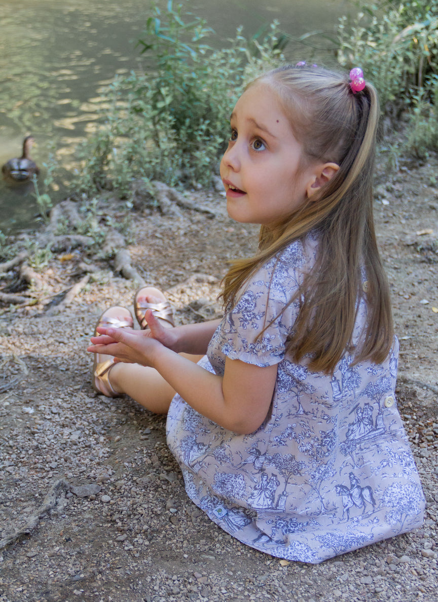 A lifestyle photograph of a young girl sitting outdoors by the edge of a pond, looking upward with an expressive, curious expression. She wears the Carmen short-sleeved dress made from light beige fabric featuring a classic grey and white toile de Jouy print, paired with metallic sandals, with greenery and a swimming duck visible in the background.