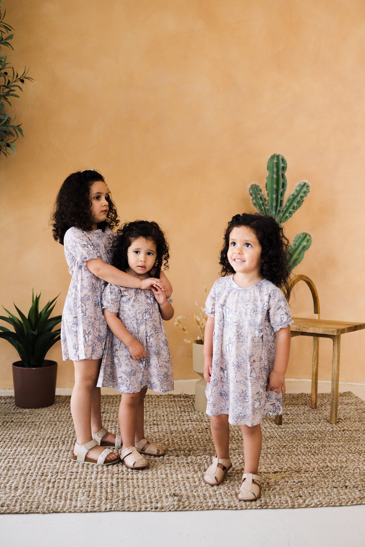 A warm studio lifestyle photograph featuring three young girls with dark curly hair posing together in matching Carmen dresses. The short-sleeved dresses feature a classic grey and white toile de Jouy print on a light beige fabric. The girls stand on a woven jute rug against a warm terracotta wall with subtle potted plants, a wooden chair, and cactus decor in the background.