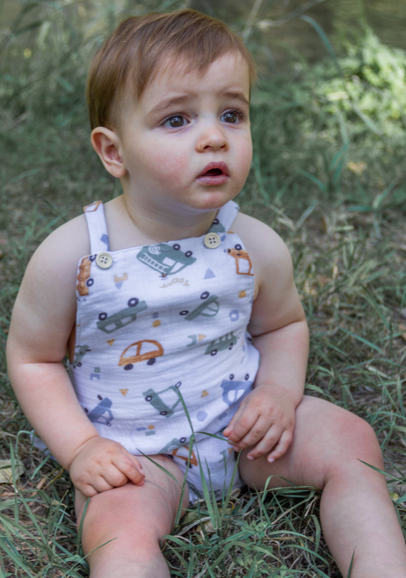 A close-up lifestyle portrait of a baby boy sitting outdoors in the grass, wearing a white cotton overall romper printed with colorful cars and vehicles. The romper features shoulder straps with small wooden buttons, and the baby is looking curiously off-camera in a natural outdoor setting.