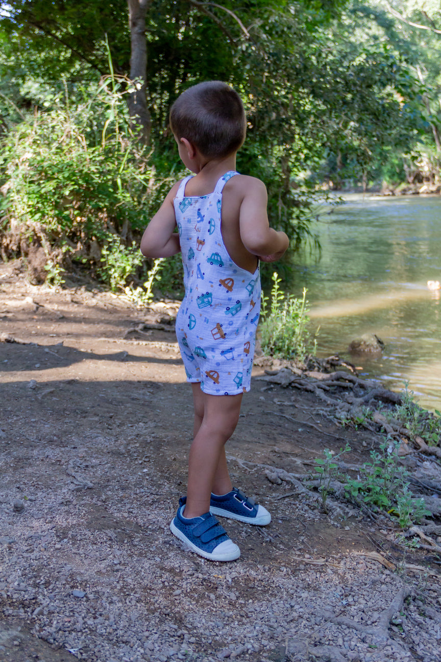 A lifestyle photograph of a young toddler boy standing outside on a dirt riverbank wearing a sleeveless light-colored overall romper patterned with colorful car and vehicle illustrations. Seen from the side and back, he wears blue sneakers while exploring near a river and lush green trees.