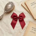 A flat lay photograph of a large, dark red grosgrain ribbon bow hair clip. The bow is centered on a cream-colored textured fabric backdrop, styled with a vintage silver-backed hairbrush and two old, weathered books with titles like "Whispers of the Heart." The scene is illuminated by soft, natural light, creating a vintage and romantic feel.