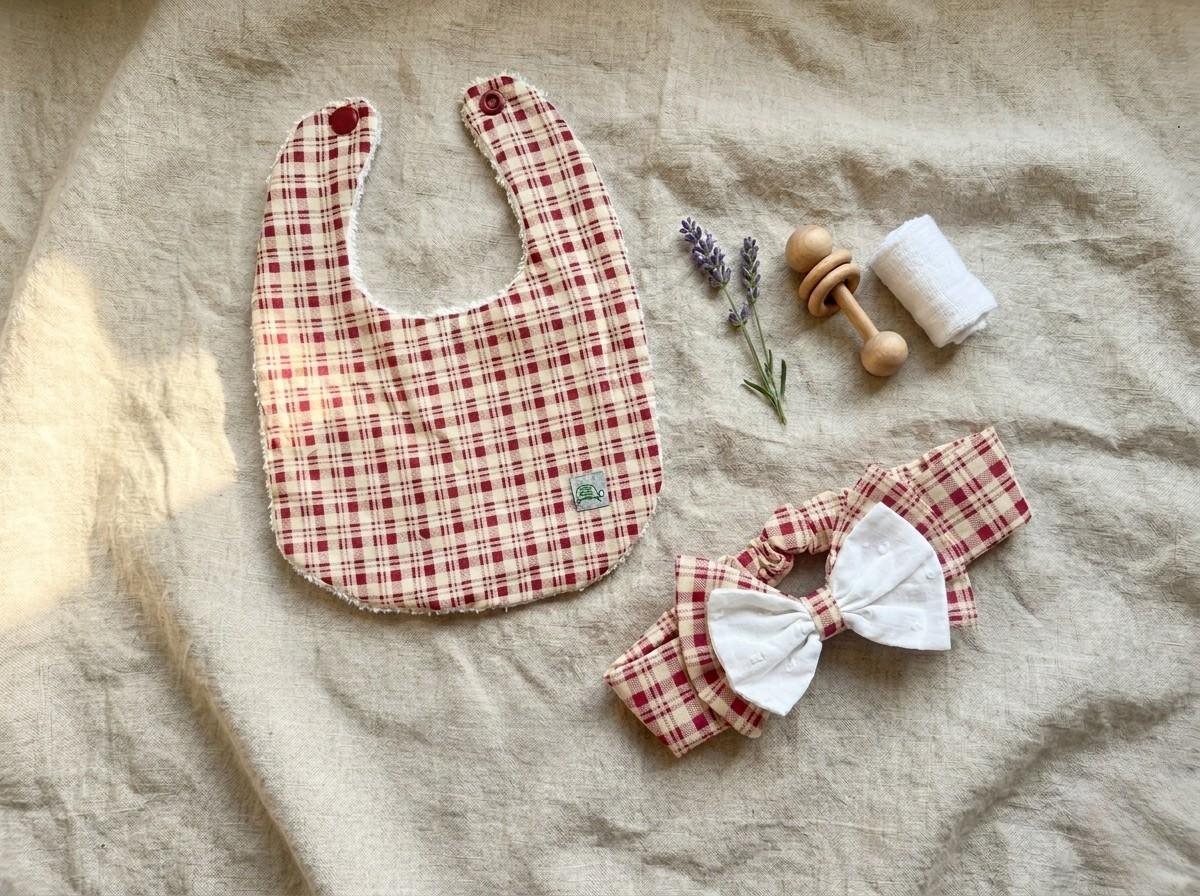 A flat lay photograph of the Cordoba set featuring a baby bib and a matching headband. Both items are made from a red and cream gingham checkered fabric. The bib has a white textured backing and red snap fasteners, while the headband features a layered double bow with a white eyelet fabric on top. They are styled on a neutral linen background alongside a wooden rattle, a sprig of dried lavender, and a small white cloth roll.