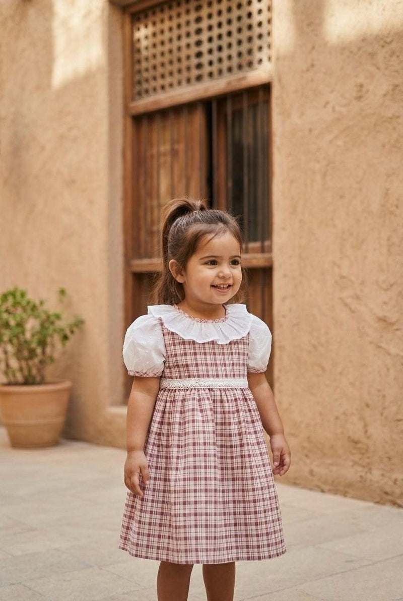 A portrait lifestyle photograph of a smiling young girl standing outside on a stone-tiled ground. She is wearing the Cordoba dress, which features a red-brown and cream checkered plaid print, white puffed short sleeves with plaid trim, a wide white ruffled collar, and a delicate white lace-style band across the waist. She has her hair styled in a high half-ponytail, against a background of a warm, sandy-toned stone wall with a rustic wooden window frame and a potted green plant.