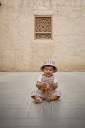 A lifestyle photograph of a smiling baby sitting barefoot and cross-legged on a grey stone-tiled floor. The baby is wearing the Cordoba set, which includes short romper overalls and a matching bucket hat in a red-brown and cream checkered plaid fabric, layered over a simple white short-sleeved t-shirt. The baby is looking down and playing with a small toy camel against a background of a warm, sandy-coloured textured stone wall with an ornate lattice window cutout.