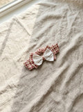A flat lay photograph of the Cordoba baby headband featuring a red and cream gingham checkered pattern. The headband is topped with a unique double bow: a larger gingham bow sits beneath a smaller, delicate white eyelet fabric bow. It is styled diagonally on a textured, neutral-toned linen fabric under soft, natural sunlight.