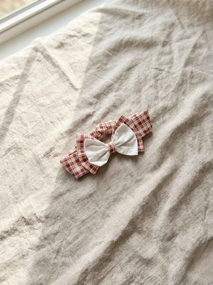 A flat lay photograph of the Cordoba baby headband featuring a red and cream gingham checkered pattern. The headband is topped with a unique double bow: a larger gingham bow sits beneath a smaller, delicate white eyelet fabric bow. It is styled diagonally on a textured, neutral-toned linen fabric under soft, natural sunlight.