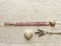 A flat lay photograph of the Cordoba pacifier clip, featuring a fabric strap with a red and cream gingham checkered pattern. The strap is gathered with an elasticated, ruffled texture and attached to a round, natural light-wood clip. It is styled on a neutral, textured cream fabric background next to a small white felt heart and several sprigs of dried lavender.