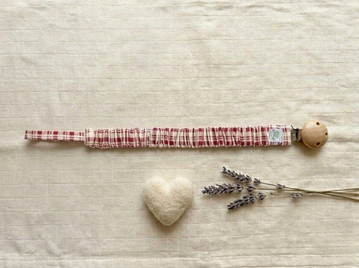 A flat lay photograph of the Cordoba pacifier clip, featuring a fabric strap with a red and cream gingham checkered pattern. The strap is gathered with an elasticated, ruffled texture and attached to a round, natural light-wood clip. It is styled on a neutral, textured cream fabric background next to a small white felt heart and several sprigs of dried lavender.