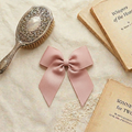 A flat lay photograph of a large, dusty pink grosgrain ribbon bow hair clip. The bow is centered on a cream-colored textured fabric backdrop, styled with a vintage silver-backed hairbrush and two old, weathered books. The scene uses soft, natural lighting to highlight the ribbed texture of the ribbon and the romantic, heirloom quality of the accessory.