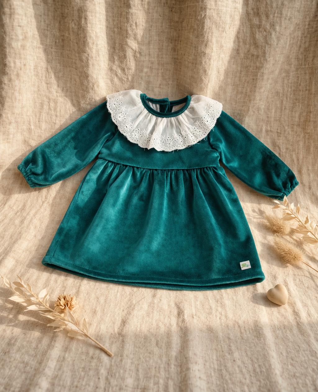A long-sleeved emerald green velvet baby dress featuring a wide white eyelet lace Peter Pan collar. The dress has a gathered waist and elasticated cuffs, displayed on a neutral linen backdrop with dried botanical accents and a small wooden heart.