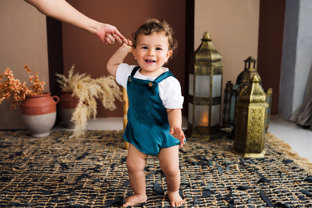 A candid studio lifestyle photograph of a cheerful toddler holding an adult's hand while taking steps on a dark woven rug. The toddler wears an emerald green velvet overall romper over a short-sleeved white t-shirt. The romper features gold button-fastened shoulder straps and elasticized leg openings. The background is styled with warm terracotta tones, ceramic pots with dried pampas grass, and decorative brass lanterns.