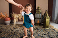 A candid studio lifestyle photograph of a cheerful toddler holding an adult's hand while taking steps on a dark woven rug. The toddler wears an emerald green velvet overall romper over a short-sleeved white t-shirt. The romper features gold button-fastened shoulder straps and elasticized leg openings. The background is styled with warm terracotta tones, ceramic pots with dried pampas grass, and decorative brass lanterns.