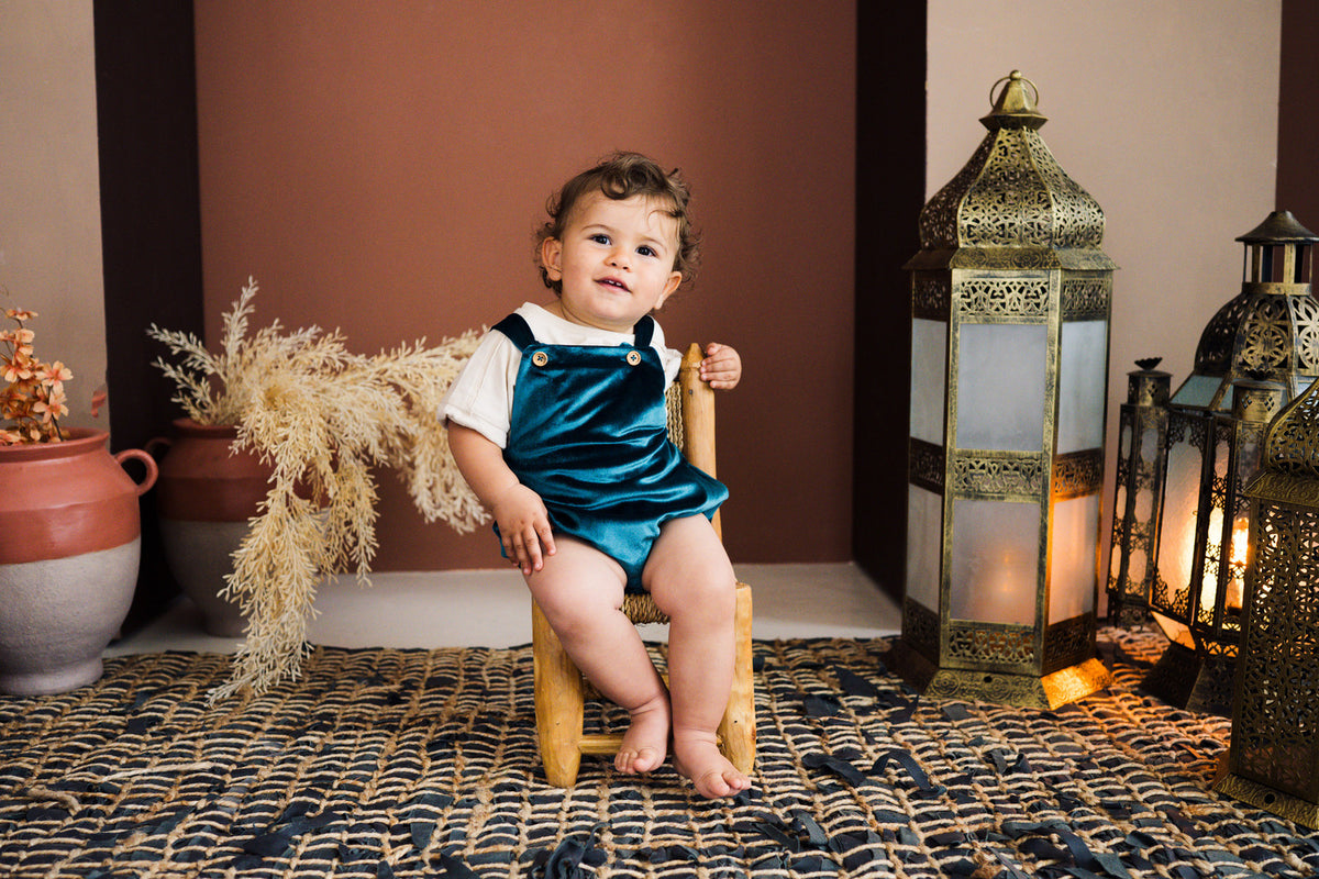 A studio lifestyle photograph of a smiling toddler sitting on a small wooden chair on a dark woven rug, wearing an emerald green velvet overall romper over a white t-shirt. The romper features shoulder straps with gold button closures and elasticized leg openings. Warm terracotta walls, dried pampas grass in ceramic pots, and traditional brass lanterns complete the backdrop.