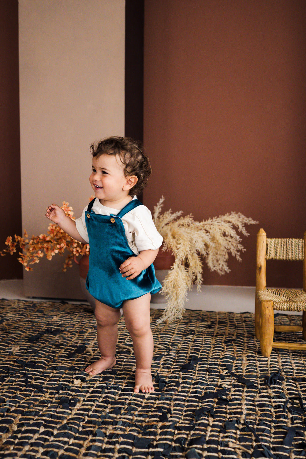 A full-length studio lifestyle photograph of a smiling toddler standing barefoot on a dark woven rug, wearing an emerald green velvet overall romper layered over a white t-shirt. The overall romper features shoulder straps with gold button details and elasticized leg openings. Warm terracotta wall panels, a small wooden chair, and dried pampas grass decorations accent the background.