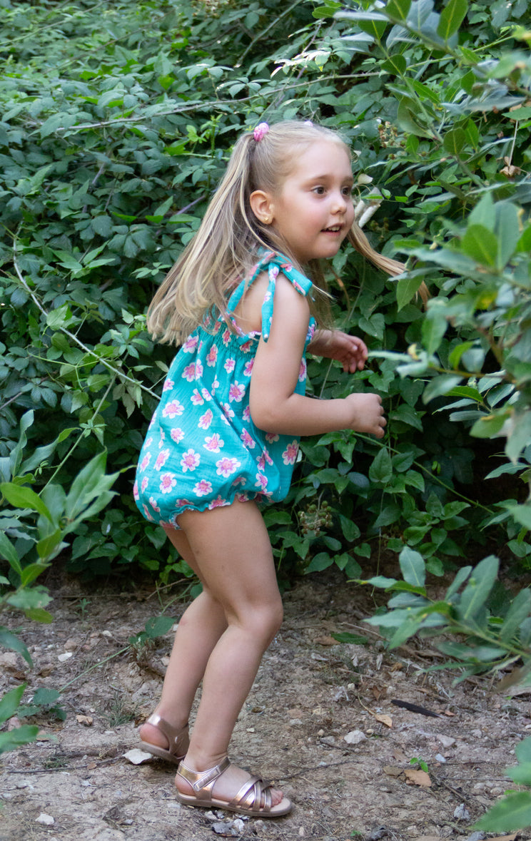 A full-length lifestyle photograph of a young blonde girl captured in a playful, dynamic stance outdoors. She is wearing the Joy romper, a teal bubble-style romper featuring a pink and white floral pattern, shoulder straps tied into bows, and a smocked bodice. She is styled with her hair in a half-up ponytail secured by a pink hair tie and is wearing metallic rose gold strappy sandals. She stands on a dirt path, leaning forward as if exploring, with a dense wall of lush green leafy bushes behind her.